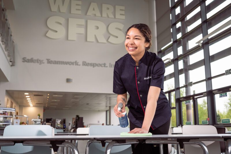 Facilities Management Photography for Sodexo at Scottish Fire and Rescue HQ, Glasgow 5 Sodexo cleaning staff member wiping down canteen tables below a "We Are SFRS" wall sign