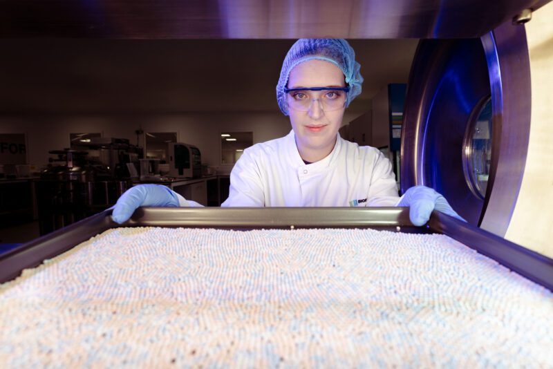 Shot Entirely In-Camera: One Freeze-Dryer Session Built Biofortuna's Exhibition Stand Hero Image 4 Scientist in cleanroom attire holding a tray of freeze-dried diagnostic beads towards camera, shot from inside an open freeze-dryer chamber with dramatic blue lighting.
