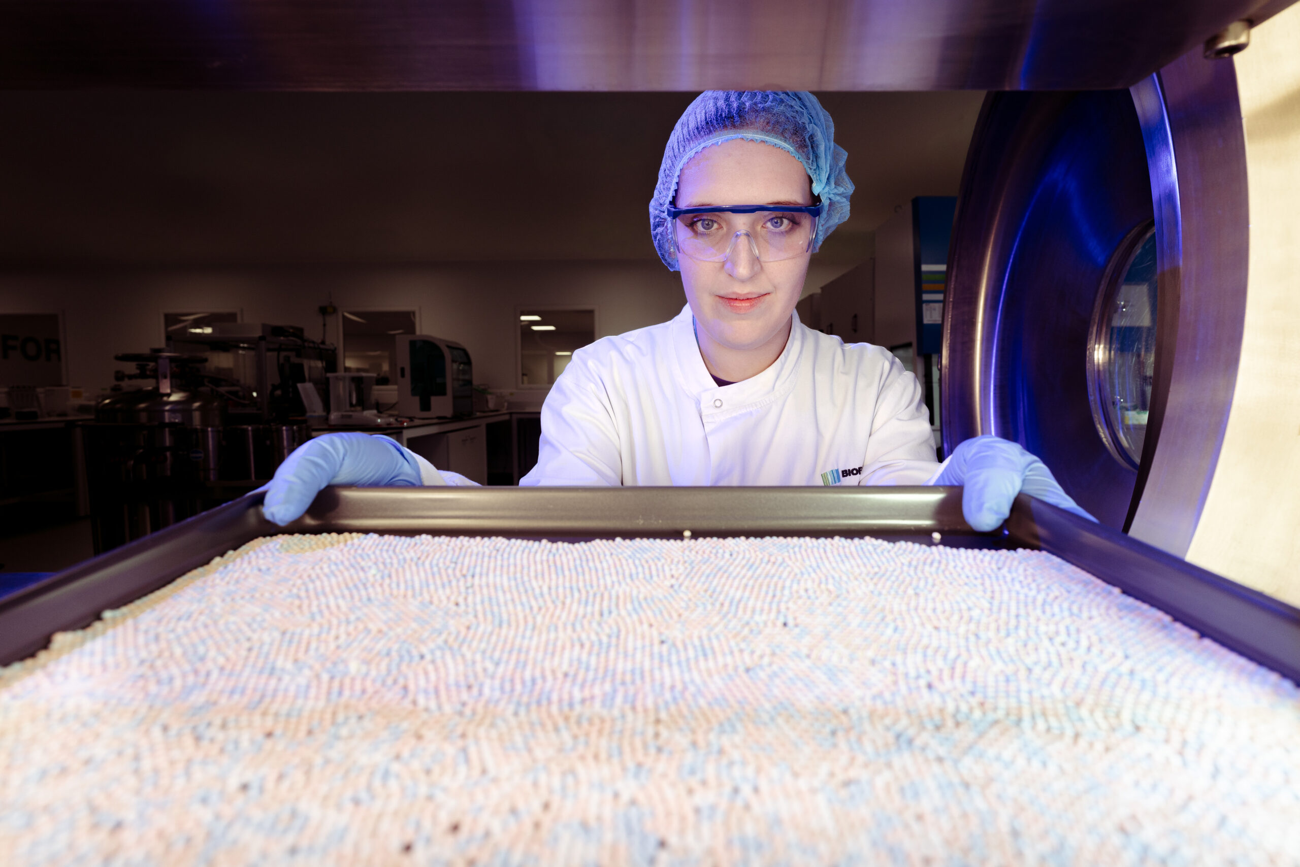 Scientist in cleanroom attire holding a tray of freeze-dried diagnostic beads towards camera, shot from inside an open freeze-dryer chamber with dramatic blue lighting.