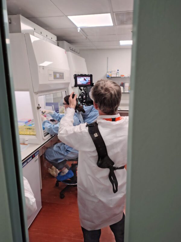 Photographing the People Behind SGS Vitrology's New Glasgow Lab 3 Photographer in PPE shooting handheld with a camera monitor rig into a biosafety cabinet, where a technician in a blue gown works, during the SGS Vitrology Glasgow shoot.