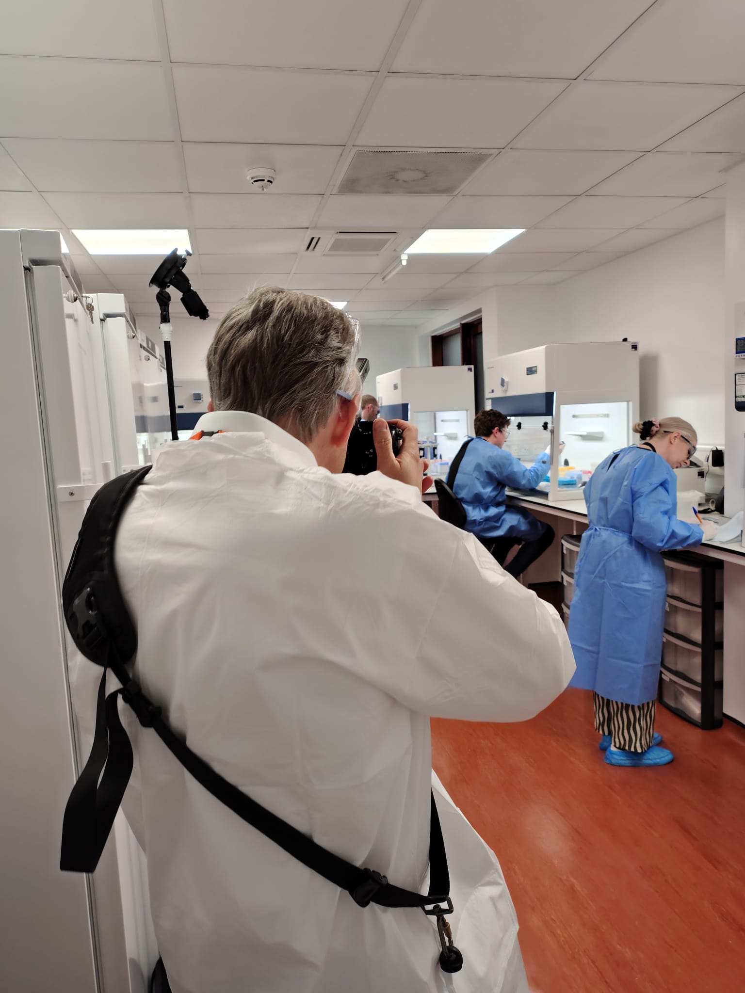 Growing a Commercial & Industrial Photography Business 7 Photographer in PPE photographing two technicians working at biosafety cabinets down a laboratory corridor, during the SGS Vitrology Glasgow shoot.