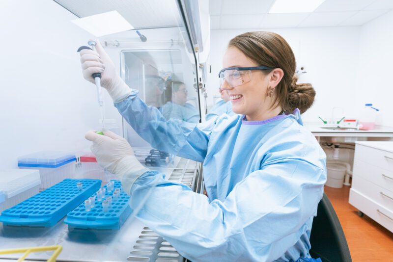 Photographing the People Behind SGS Vitrology's New Glasgow Lab 1 Female scientist in PPE pipetting a sample inside a biosafety cabinet at SGS Vitrology's Glasgow laboratory, with a colleague reflected in the cabinet glass.