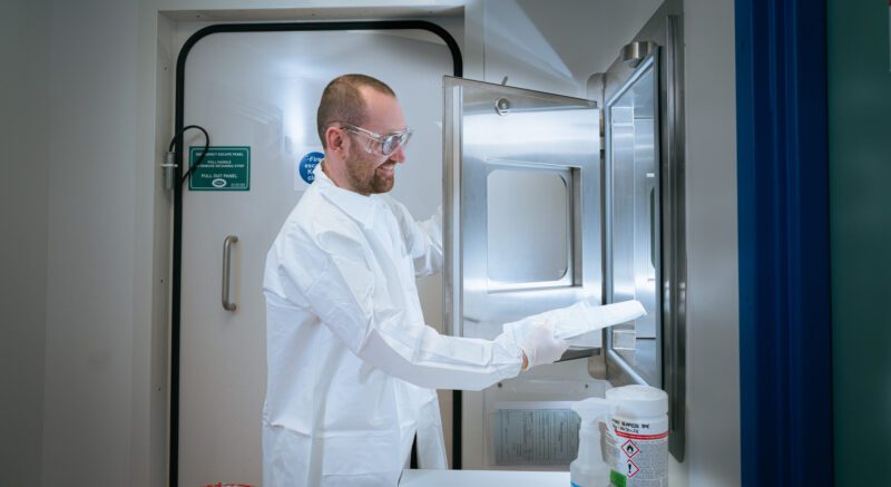 Photographing the People Behind SGS Vitrology's New Glasgow Lab 11 Technician in full PPE smiling while placing a sealed sample package into a stainless steel pass-through sterilisation hatch at SGS Vitrology's Glasgow lab.