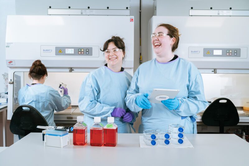 Photographing the People Behind SGS Vitrology's New Glasgow Lab 7 Two SGS Vitrology scientists in blue PPE laughing together beside cell culture bottles and BioMAT safety cabinets in the Glasgow laboratory.