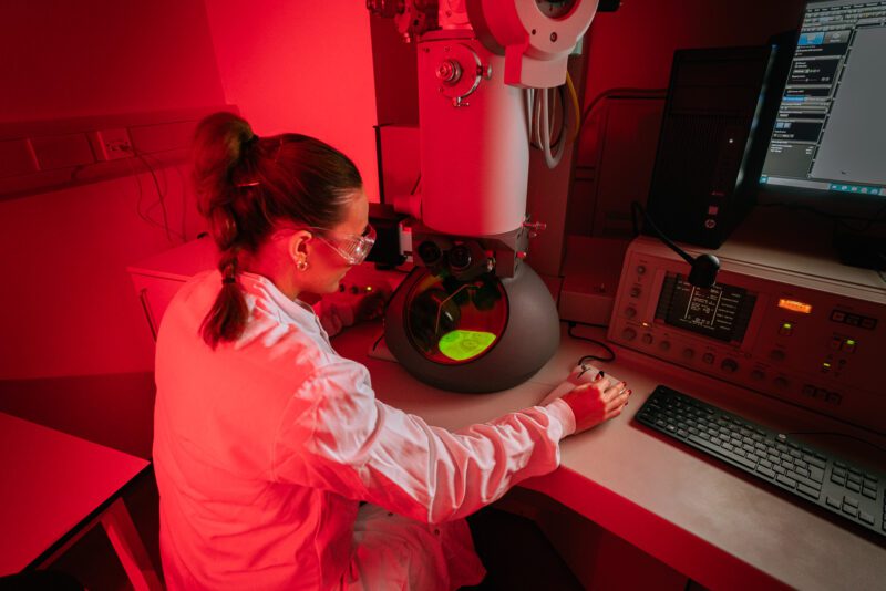 Scientist in a lab coat and safety glasses operating an electron microscope under red safelight at SGS Vitrology's Glasgow laboratory.