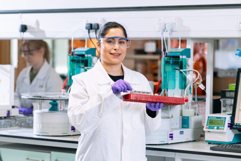 Photographing the People Behind SGS Vitrology's New Glasgow Lab 12 Analyst in a white lab coat and purple gloves smiling while holding a tray of sample vials in SGS Vitrology's Glasgow laboratory.