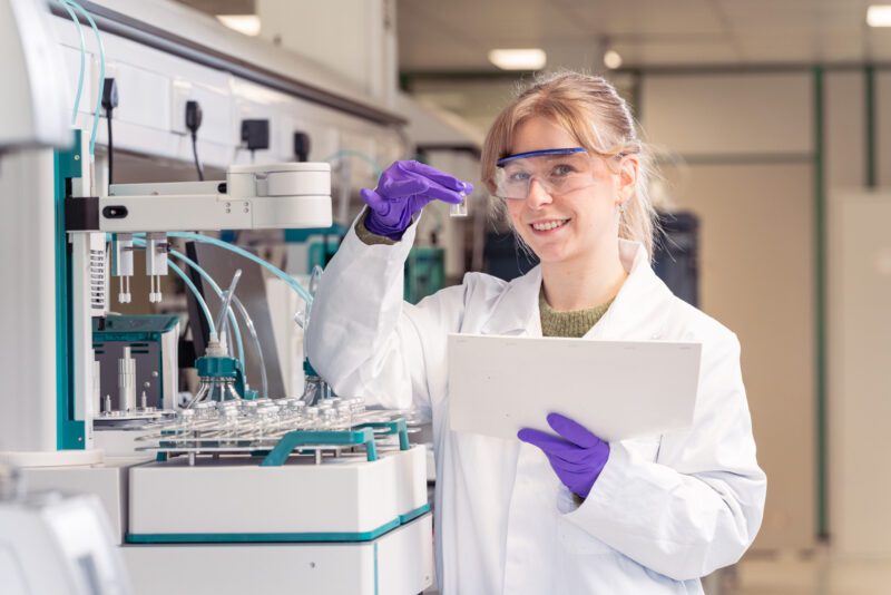 Photographing the People Behind SGS Vitrology's New Glasgow Lab 13 Analyst examining a sample vial beside a gas chromatography instrument while holding a clipboard at SGS Vitrology's Glasgow laboratory.
