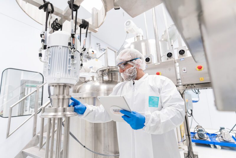 Photographing the People Behind SGS Vitrology's New Glasgow Lab 2 Technician in full cleanroom gown and hairnet checking equipment on a stainless steel processing vessel while holding a tablet at SGS Vitrology's Glasgow manufacturing suite.