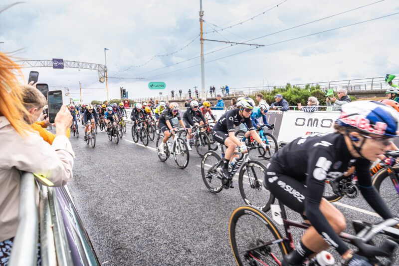Shooting OMODA's Brand Activation at the Tour of Britain Women's, Blackpool 9 A tightly packed group of women's cyclists, including a rider in an orange helmet, sprints along the seafront course watched by spectators with phones raised