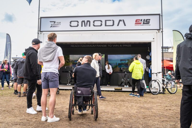 Shooting OMODA's Brand Activation at the Tour of Britain Women's, Blackpool 8 A man using a sports wheelchair watches large screens showing the OMODA simulated cycling course inside the brand activation unit, surrounded by other visitors