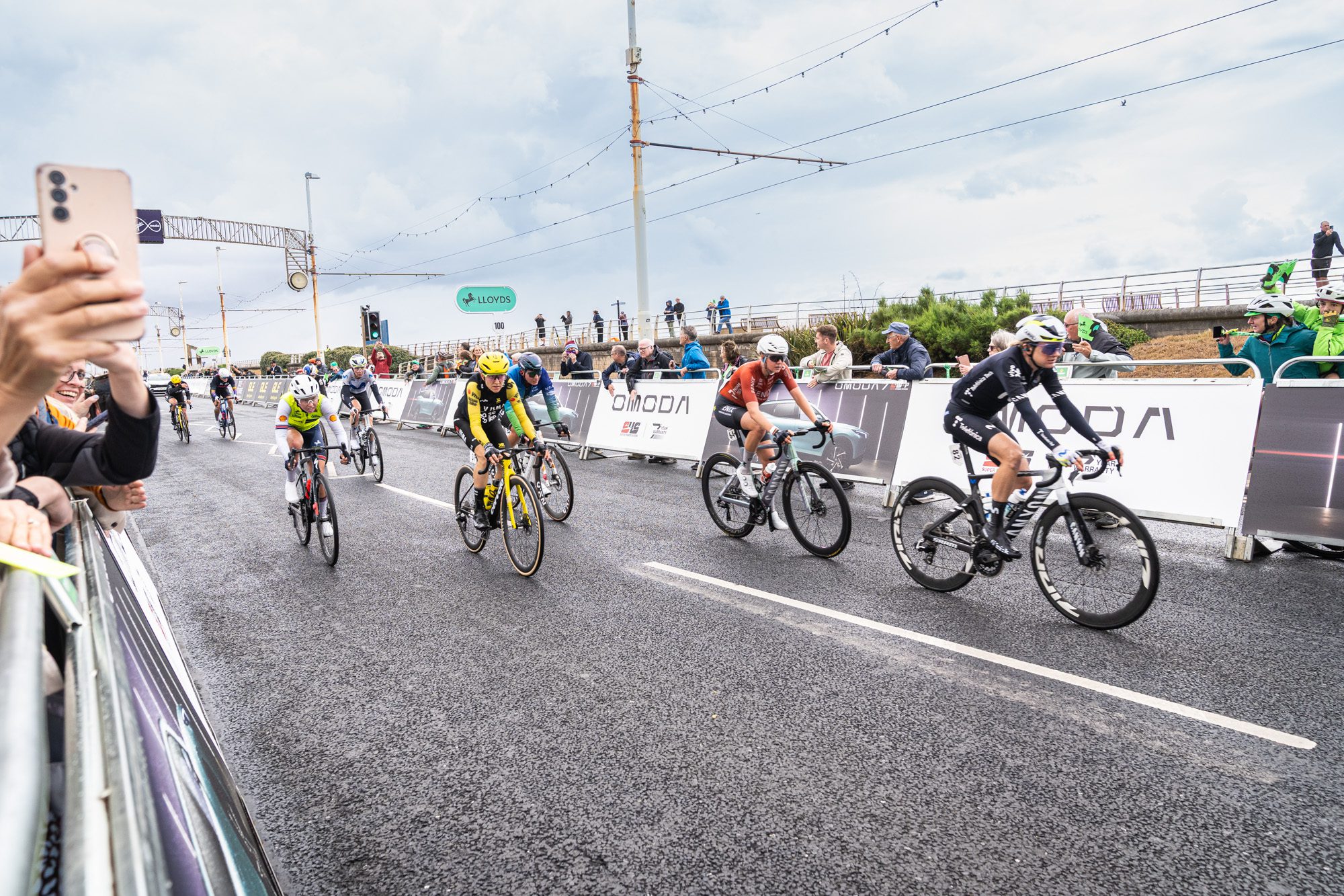 Shooting OMODA's Brand Activation at the Tour of Britain Women's, Blackpool 11 A breakaway group of women's road cyclists, led by a rider in a yellow helmet, races along Blackpool promenade past OMODA branded barriers near the finish line