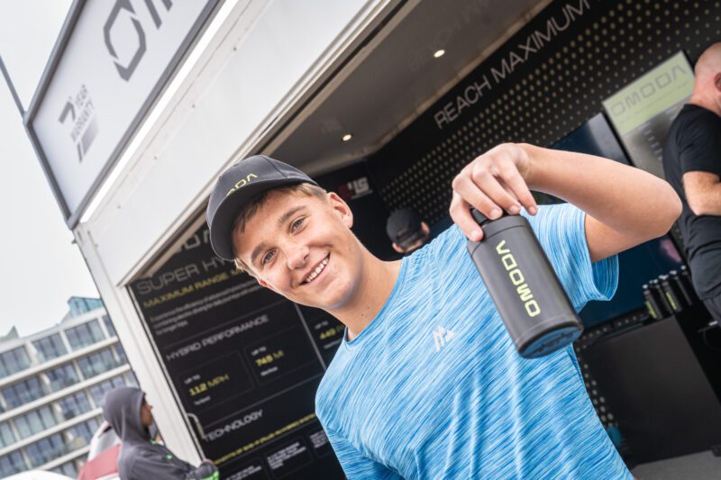 Shooting OMODA's Brand Activation at the Tour of Britain Women's, Blackpool 2 A smiling teenager in an OMODA cap holds up a black OMODA branded water bottle inside the brand activation unit