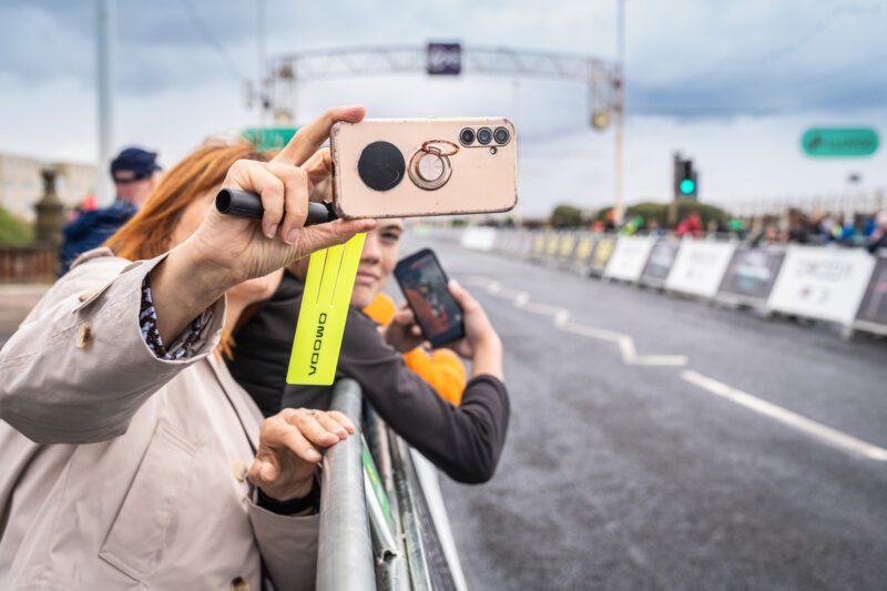 Shooting OMODA's Brand Activation at the Tour of Britain Women's, Blackpool 6 Close-up of a spectator's hands holding a rose gold phone with a pop-socket ring, filming the race along the barriers in Blackpool