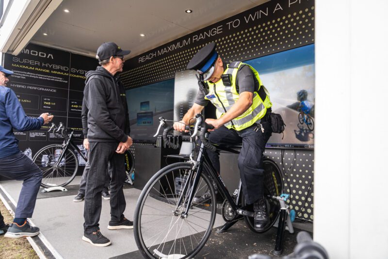 Shooting OMODA's Brand Activation at the Tour of Britain Women's, Blackpool 7 A police officer in uniform and hi-vis rides the OMODA simulated cycling trainer inside the brand activation unit, watched by a man in an OMODA cap beside the Super Hybrid System information panel