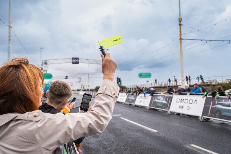 Shooting OMODA's Brand Activation at the Tour of Britain Women's, Blackpool 5 A family in green Lloyds Tour of Britain caps takes a selfie together in front of the results screen at the Blackpool Event Village, under the "get riding, Britain" banner